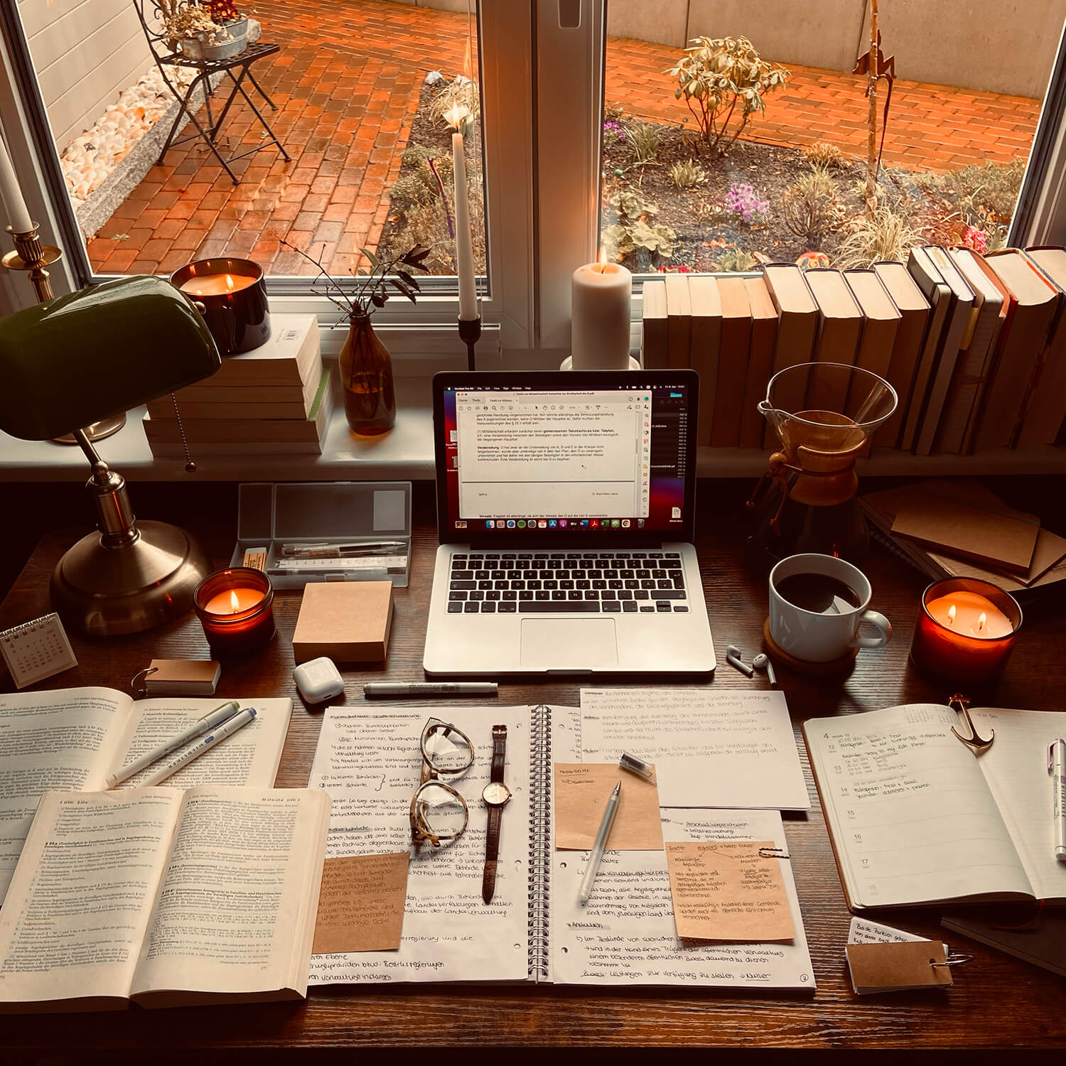 “Cozy student study desk with an open laptop, books, handwritten notes, candles, and coffee arranged by a window on a rainy day.”